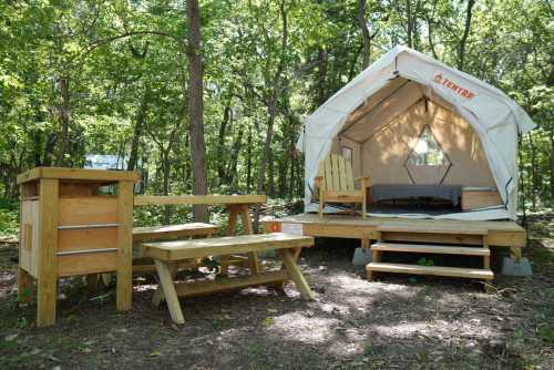 A cozy glamping tent with wooden furniture, including a picnic table and chairs, set in a lush forest.