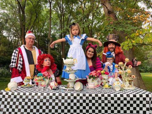 A whimsical tea party scene with characters dressed as Alice, the Mad Hatter, and the Queen of Hearts in a forest setting.