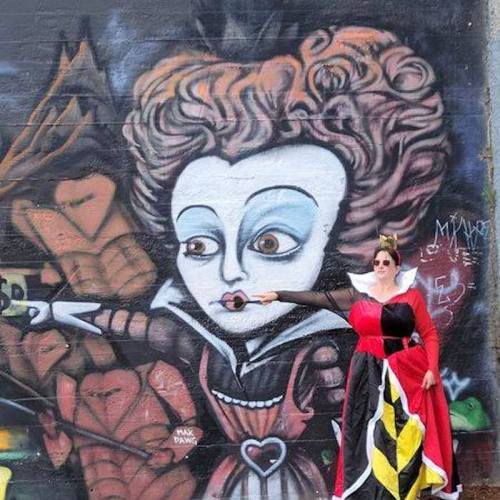 A woman in a queen costume poses in front of a colorful mural featuring a stylized queen character.