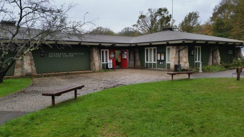 A nature center building with stone accents, surrounded by grass and trees, on a cloudy day. Benches are visible in front.