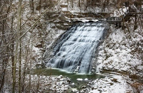 A snowy waterfall cascades down rocky cliffs, surrounded by bare trees and a serene winter landscape.