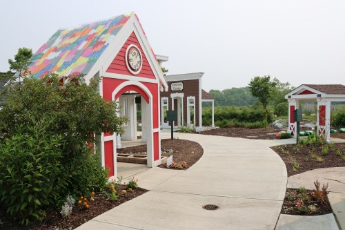 Colorful small buildings with a garden path, surrounded by greenery and flowers on a sunny day.