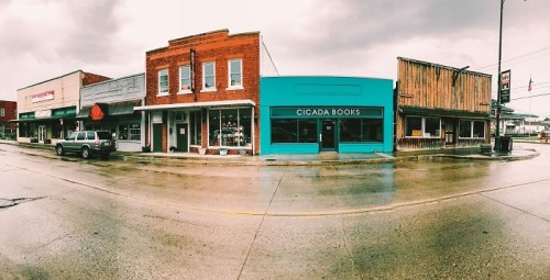A colorful street scene featuring shops, including a bright blue store named Cicada Books, on a rainy day.