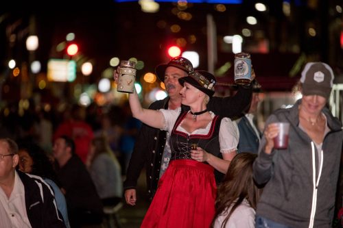 A woman in traditional attire raises a beer stein, celebrating at a lively outdoor event with a crowd in the background.