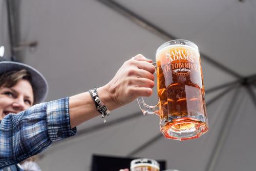 A person raises a large mug of amber beer at a festival, wearing a plaid shirt and a hat.
