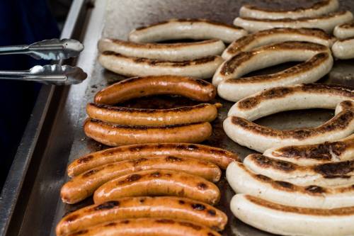 Grilled sausages on a hot griddle, featuring a mix of brown and white varieties. Tongs are placed nearby.
