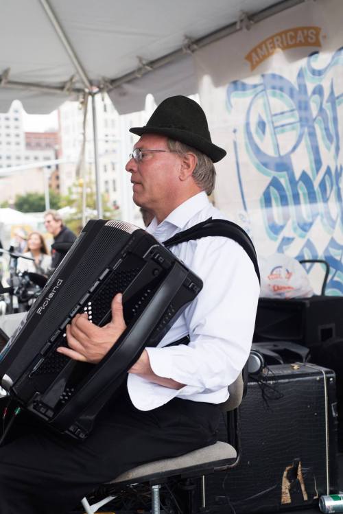 A musician in a black hat plays an accordion at an outdoor event, with a banner in the background.