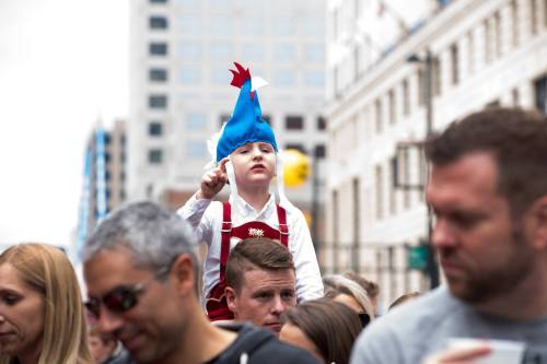 A child in a blue chicken hat sits on an adult's shoulders, pointing excitedly at a parade. Crowds are visible in the background.