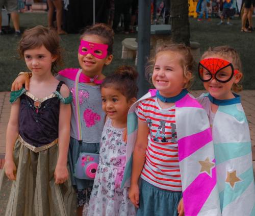 A group of four young girls in colorful costumes, smiling together at an outdoor event.