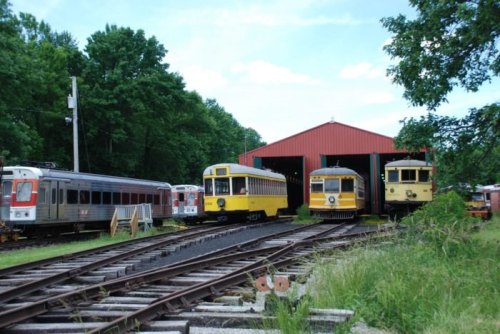 Three vintage trains in front of a red barn, surrounded by greenery and railway tracks under a blue sky.