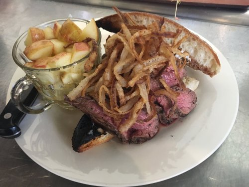 A plate featuring a beef sandwich topped with crispy onions, served with a side of diced apples in a glass bowl.