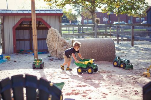 A child plays with a toy tractor in a sandy playground, surrounded by other toy vehicles and a play structure.