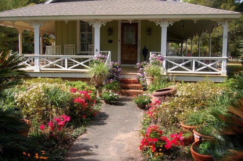 A charming house with a porch, surrounded by colorful flower beds and lush greenery.