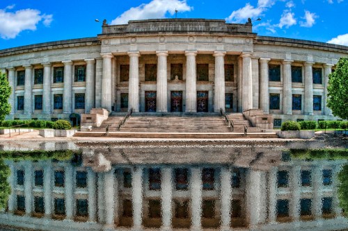A grand building with columns and steps, reflecting in a calm body of water under a blue sky with clouds.