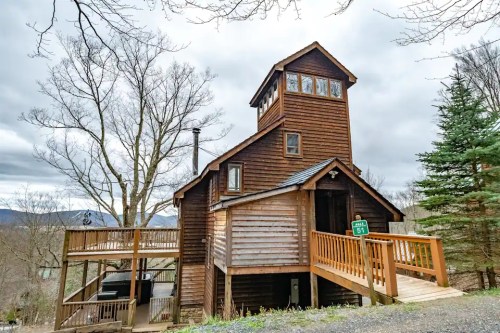A rustic wooden house with a tower, surrounded by trees and mountains under a cloudy sky.