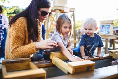 A woman and two children play together at a wooden water table, exploring with small objects in a sunny outdoor setting.