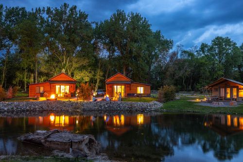 Two cozy cabins by a serene pond, illuminated at dusk, surrounded by lush trees and a cloudy sky.
