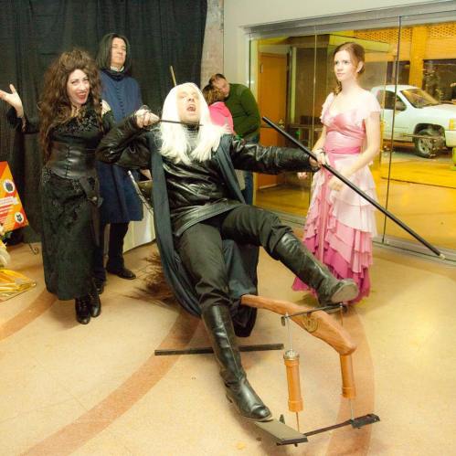 A group of four people in costumes, with one dramatically posing on a prop, while others look on in playful amusement.