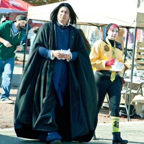A man in a long black cloak walks with a woman in a yellow shirt and tie, both at an outdoor event.