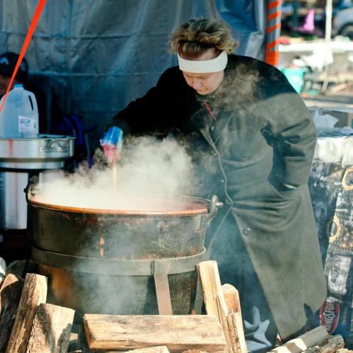 A person in a black coat stirs a large pot over a fire, surrounded by steam and wooden logs.