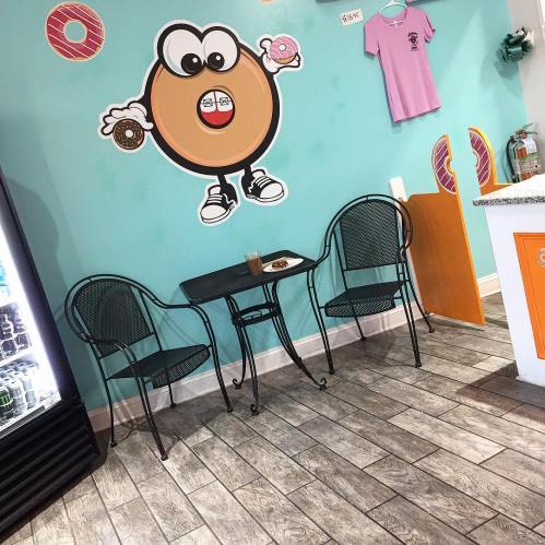 A cozy cafe corner with a cartoon donut on the wall, a small table, two chairs, and a drink in the foreground.
