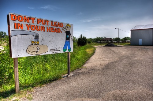 A roadside sign warns against lead exposure, featuring a cartoon character and a message about safety. Green grass and a building are in the background.