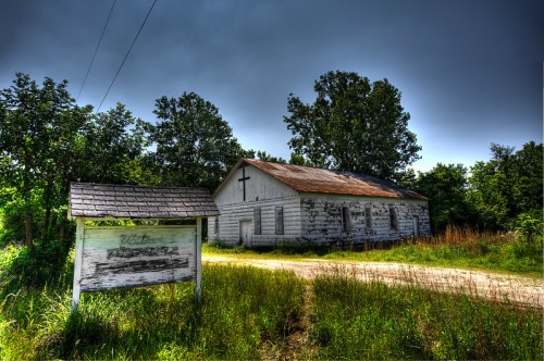 Abandoned church building with a cross, surrounded by overgrown grass and trees under a moody sky.