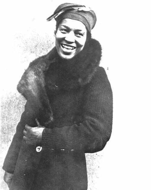 A smiling woman in a fur-collared coat and headscarf, posing against a plain background.