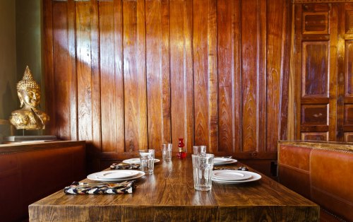 A wooden dining table set with plates and glasses, against a warm wooden wall and a decorative Buddha statue.