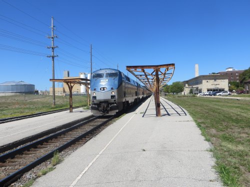 A train approaches a small station with metal canopies, set against a clear blue sky and grassy landscape.