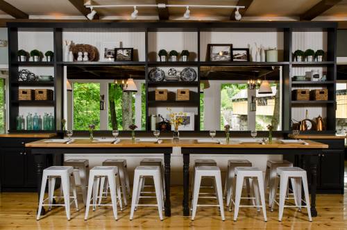 A modern kitchen with a long wooden table, white stools, and shelves displaying decorative items and plants.