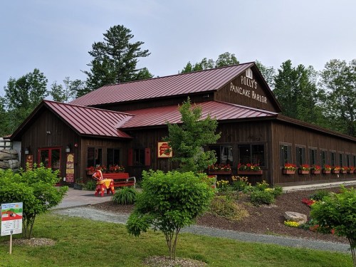 A rustic wooden building with a red metal roof, labeled "Polly's Pancake Parlor," surrounded by greenery and flowers.