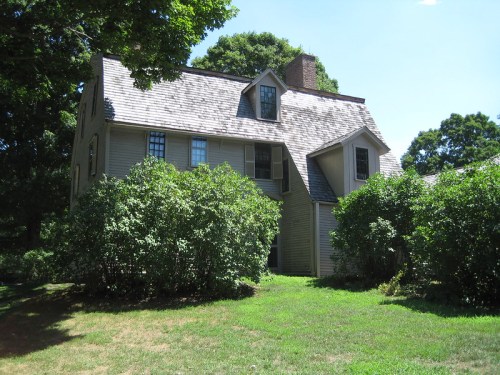 A large, two-story house with a steep roof, surrounded by green bushes and trees on a sunny day.
