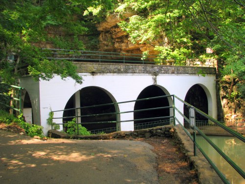 A white structure with arched openings, surrounded by greenery and a path leading to a calm waterway.