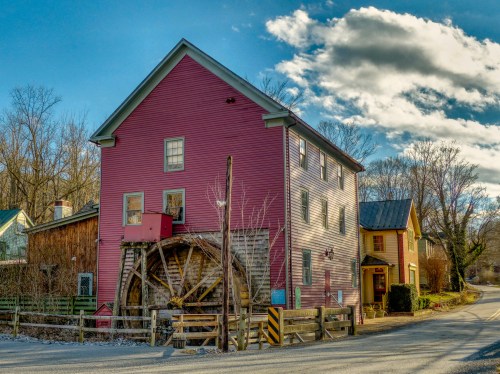A colorful old mill with a water wheel, surrounded by trees and a clear blue sky, along a quiet road.