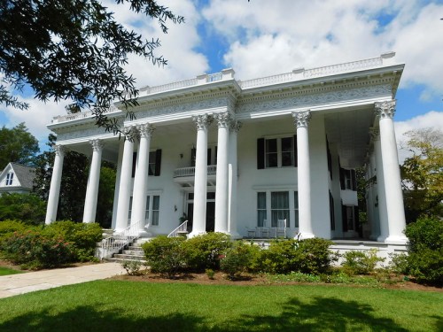 A grand white mansion with tall columns, surrounded by greenery and under a partly cloudy sky.