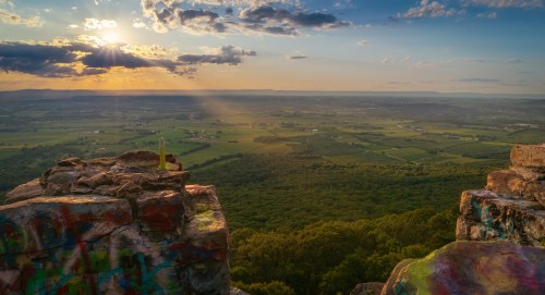 A scenic view from a rocky ledge overlooking lush green valleys and distant mountains at sunset.