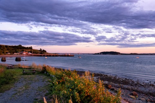 A serene coastal scene at sunset, featuring calm waters, a distant pier, and dramatic clouds overhead.