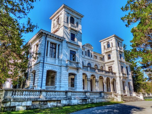 A grand, white mansion with multiple towers and balconies, surrounded by trees under a clear blue sky.