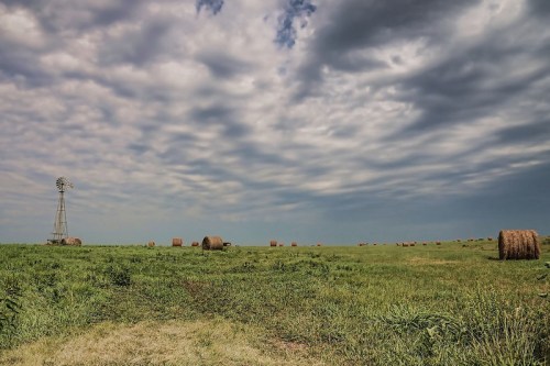 A rural landscape featuring a windmill, hay bales, and a cloudy sky over a green field.