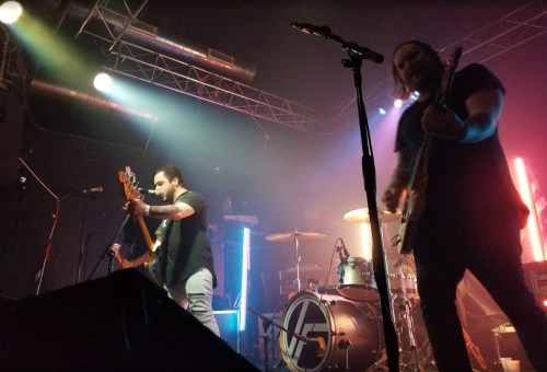 Two musicians perform on stage with guitars, illuminated by colorful lights and a drum set in the background.