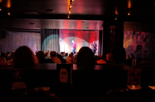 A dimly lit comedy club with a stage, featuring performers and an audience seated at tables.