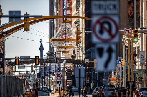 A busy city street with traffic signs, a large chandelier overhead, and pedestrians crossing in the foreground.