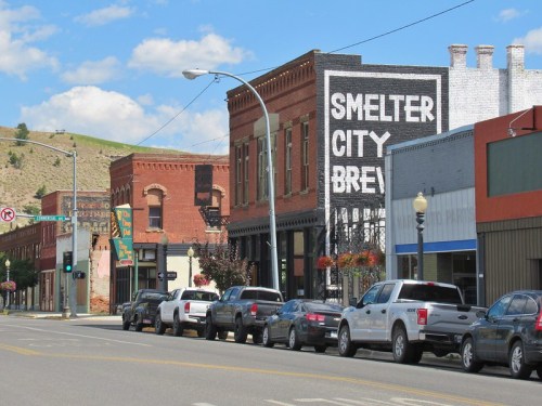 A street view of a small town featuring brick buildings, a brewery sign, and parked cars along the road.