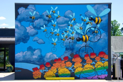 Colorful mural featuring bees on bicycles flying among clouds and vibrant flowers.