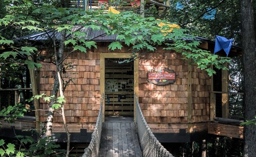 A wooden treehouse with a bridge, surrounded by green leaves and trees, featuring a sign on the front.