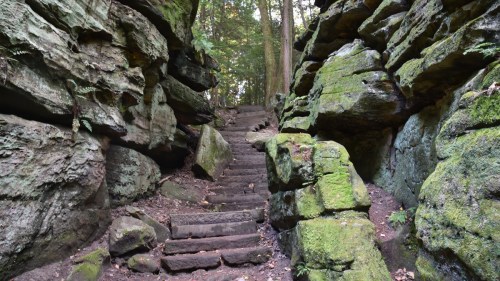 A moss-covered stone staircase leads through a narrow, rocky passage in a forested area.