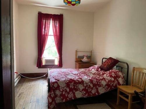 A simple bedroom with a bed covered in a floral red quilt, a chair, and a window with red curtains.