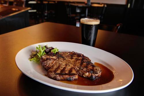 A grilled steak on a white plate with a side salad, accompanied by a glass of dark beer on a black table.
