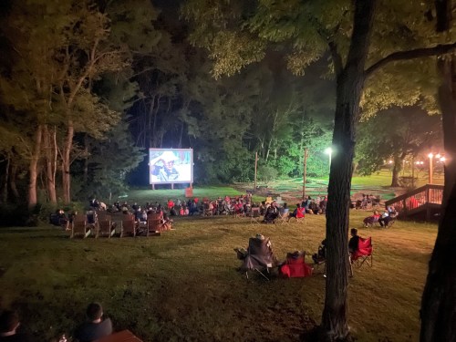 A crowd sits on a grassy area at night, watching a movie on a large outdoor screen surrounded by trees.